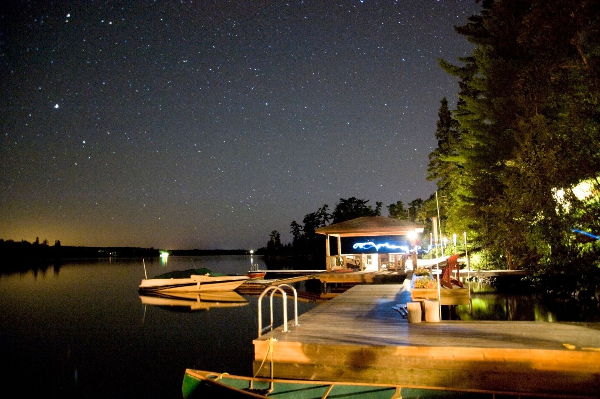 Nighttime lakeside scene with dock, building, boat, trees, and starry sky.