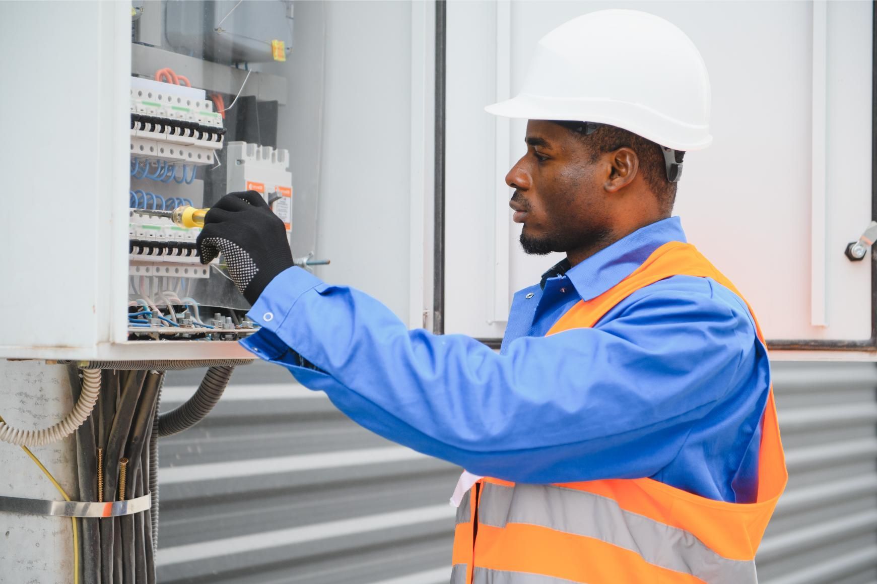 Electrician in blue workwear and orange vest working on electrical panel outdoors.