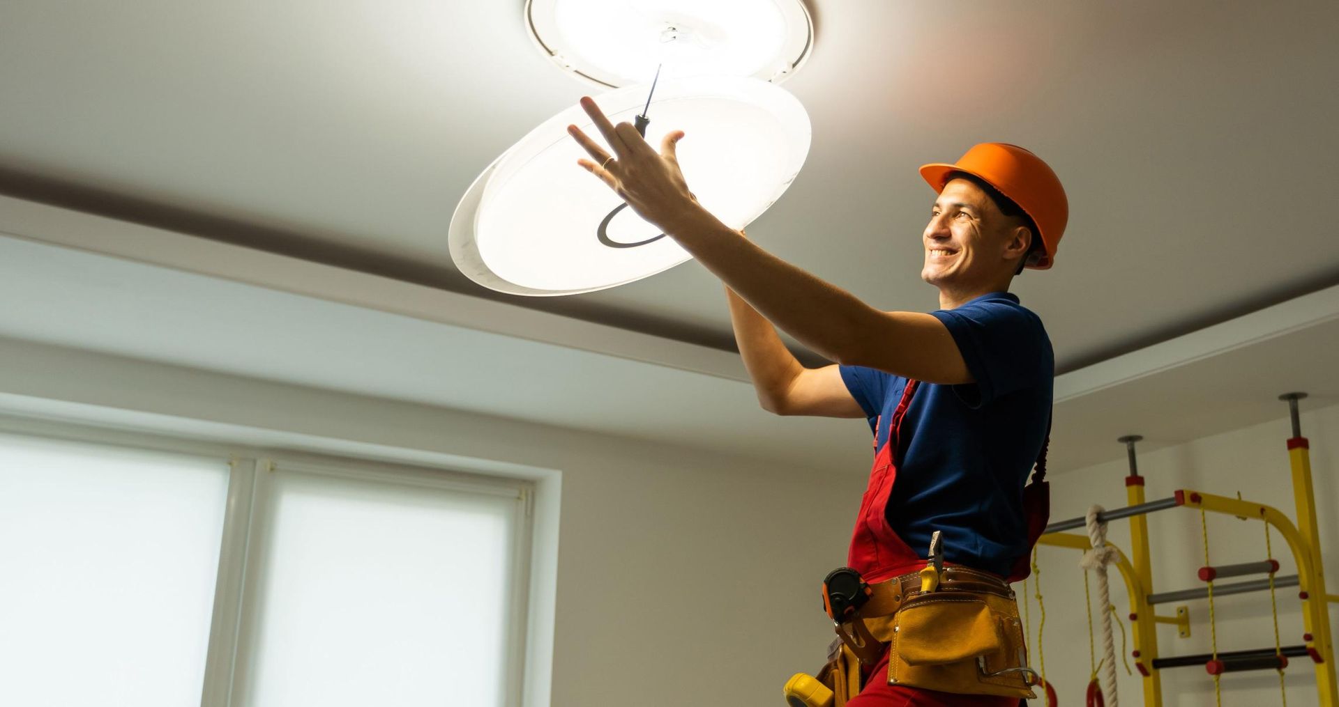 Electrician installing ceiling light in a room, wearing orange hard hat and red overalls.