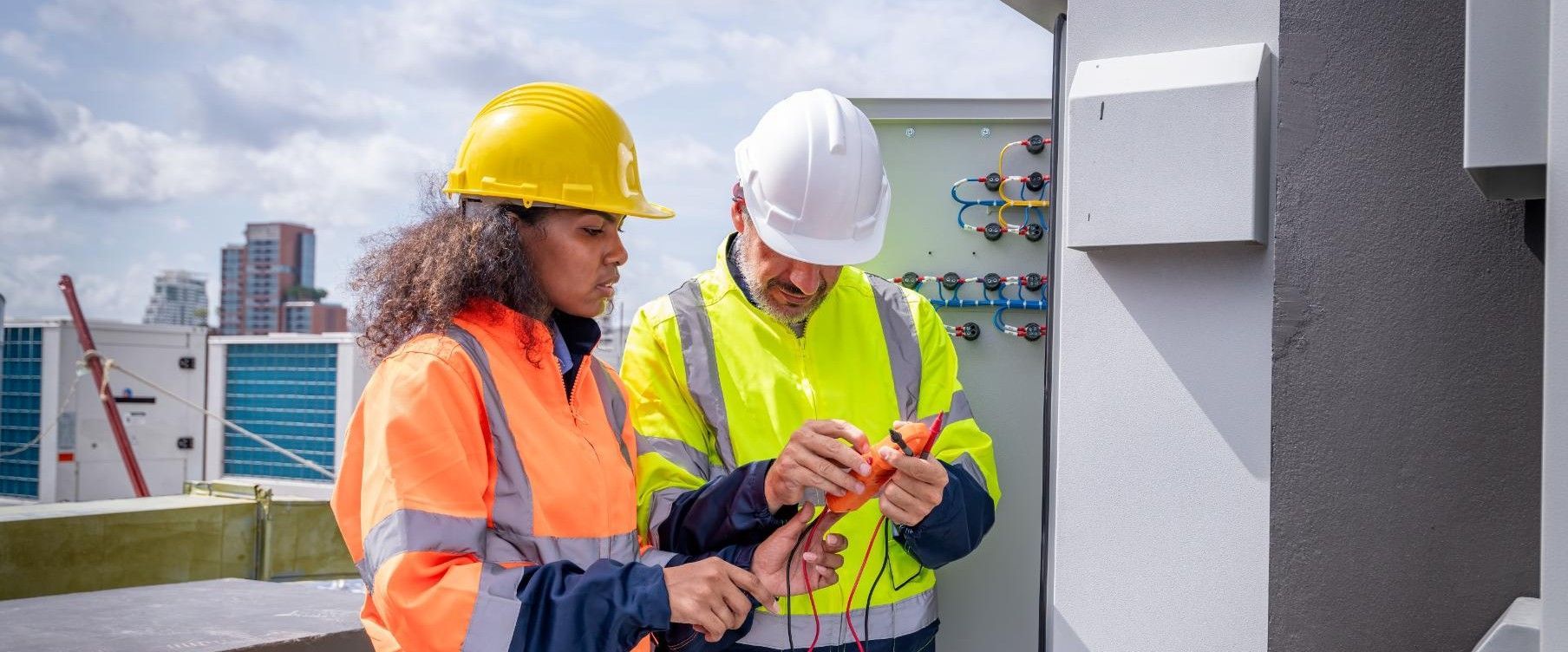 Two people in safety vests and hard hats examining electrical equipment on a rooftop.