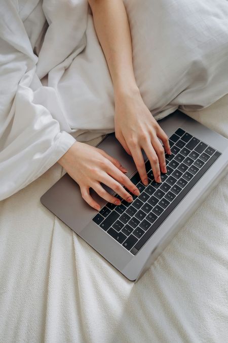 Hands typing on a laptop in bed with white bedding.