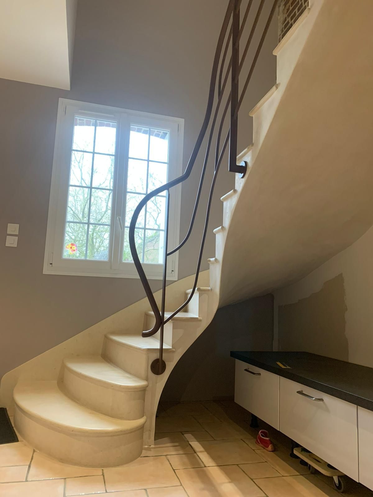Escalier courbé en pierre blanche avec une rampe en métal foncé, près d'une fenêtre et d'armoires.