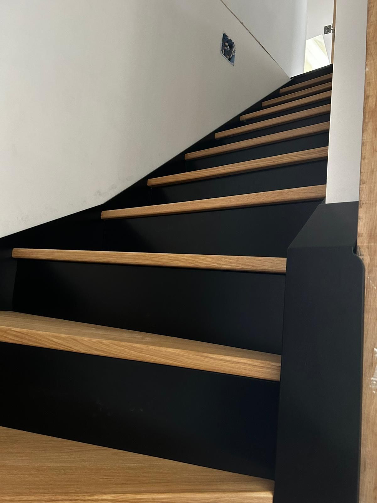 Escalier noir et bois montant, contre un mur blanc.