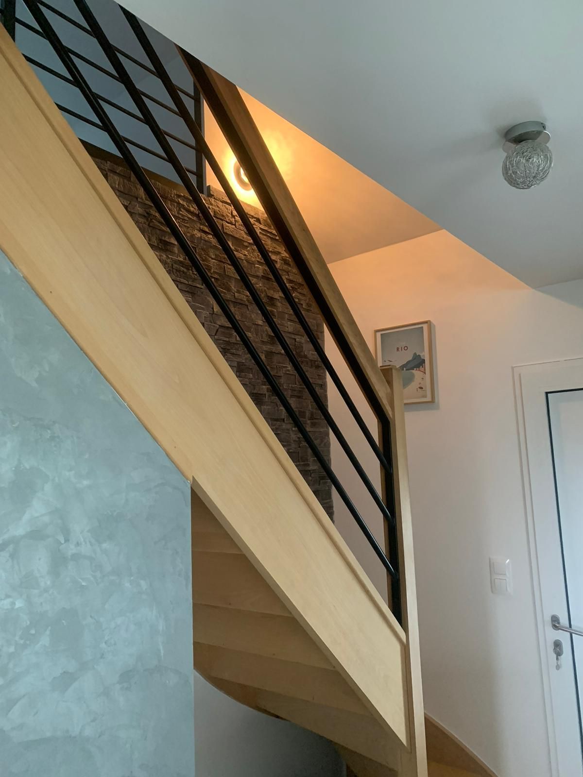 Escalier en bois avec rampe en métal noir menant vers le haut. Bois clair et mur gris visibles.