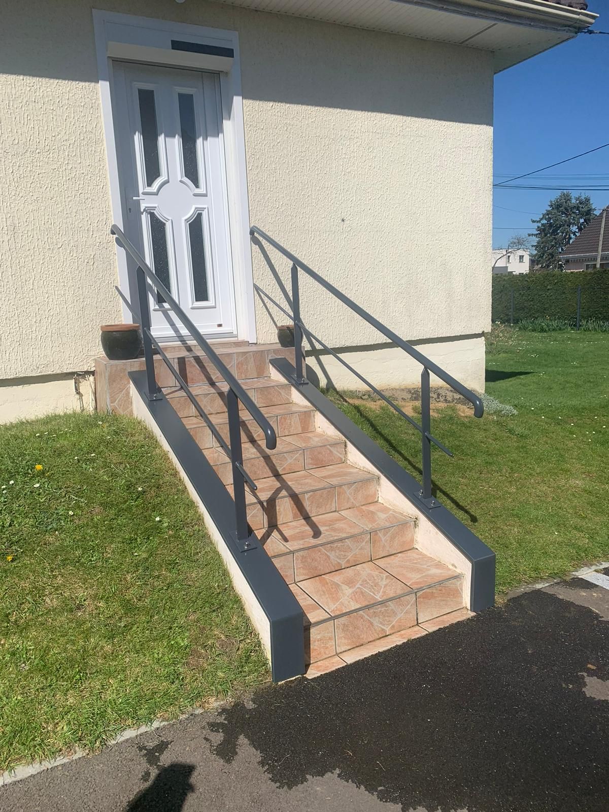 Rampe métallique grise le long des marches menant à une porte blanche. Les marches sont couleur brique et le bâtiment beige clair.