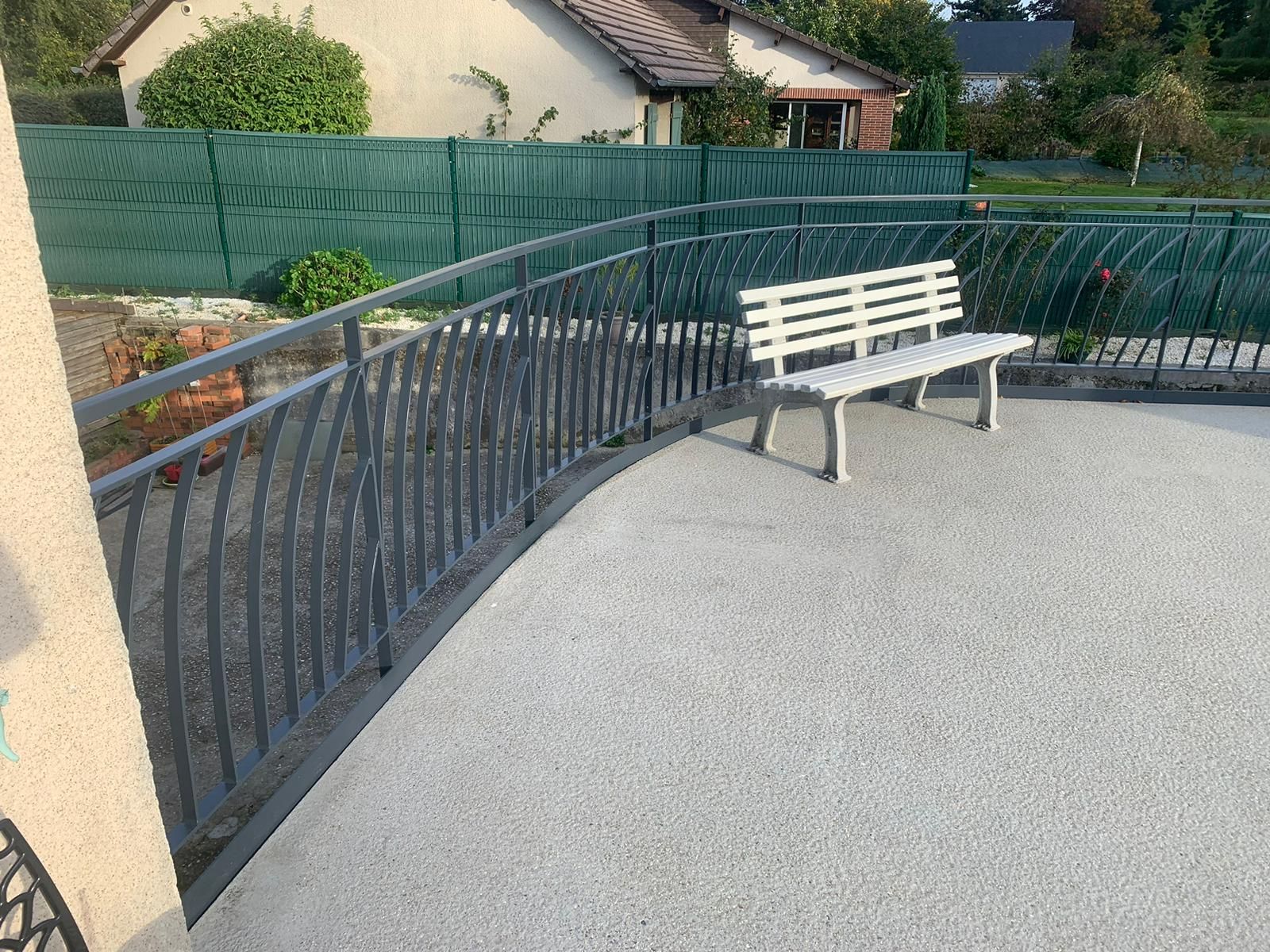 Une balustrade en métal gris s'enroule autour d'un patio avec un banc blanc, contre une clôture verte et des maisons.