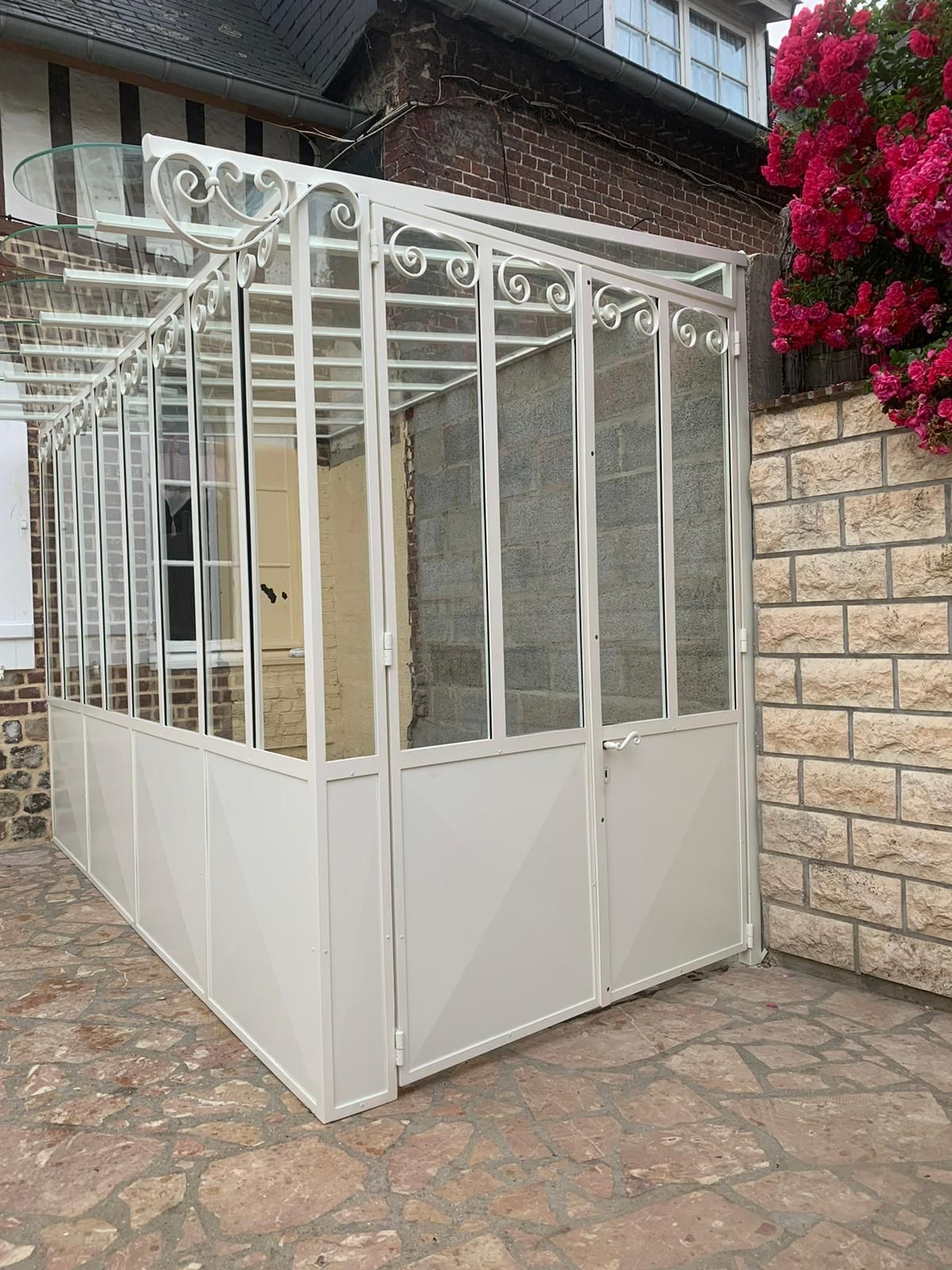 Véranda en verre blanc et métal attachée à un bâtiment en briques, vue extérieure.