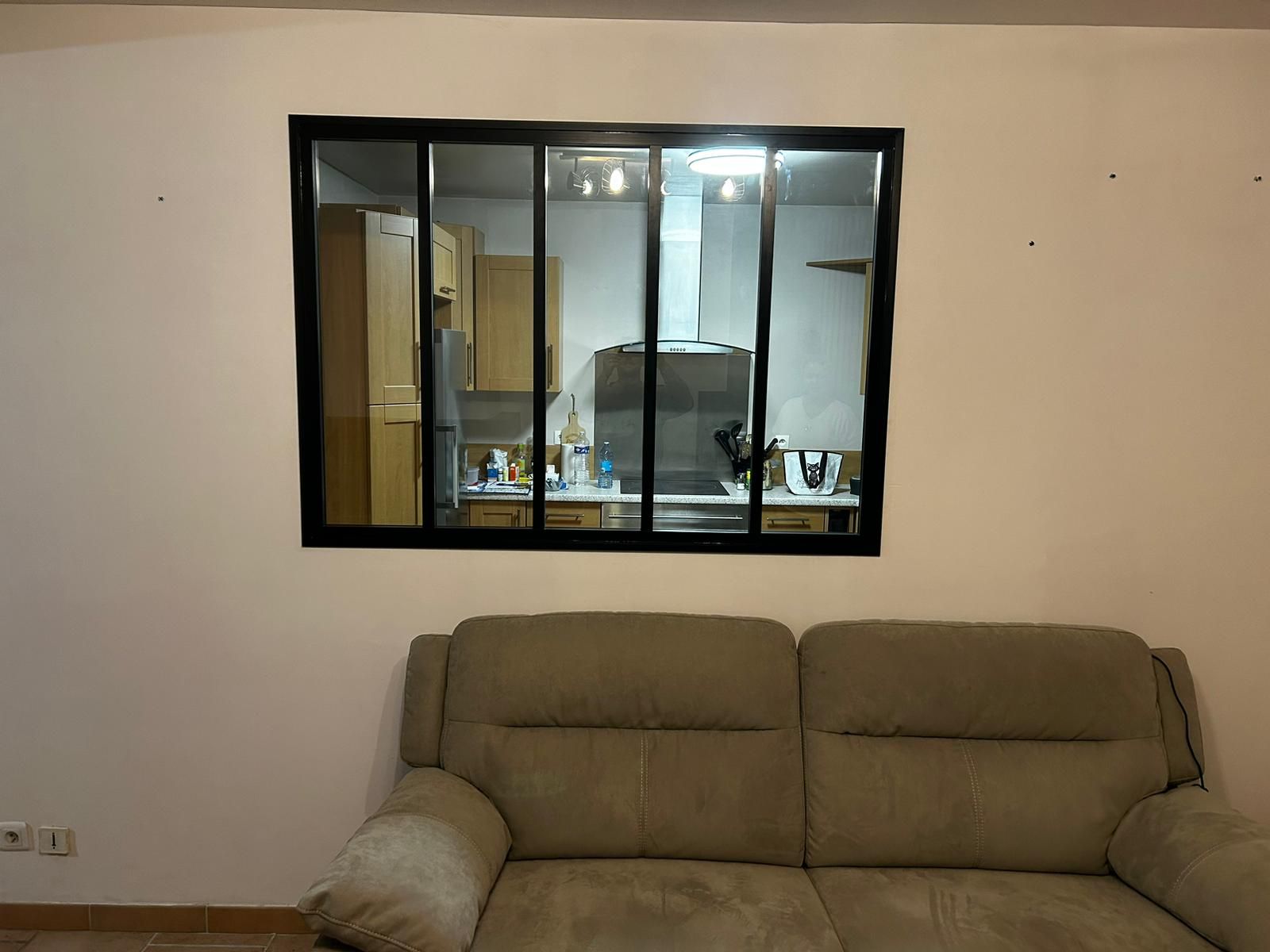 Un canapé beige trône sous une fenêtre à cadre noir donnant sur une cuisine. Les murs sont blancs.