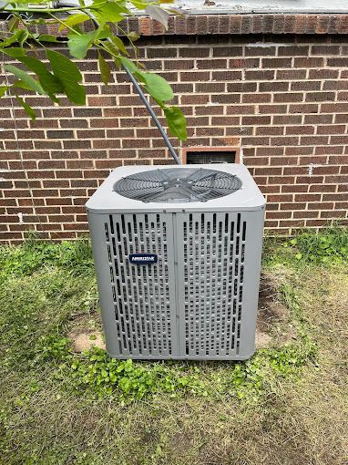 A gray air conditioner is sitting in the grass in front of a brick wall.