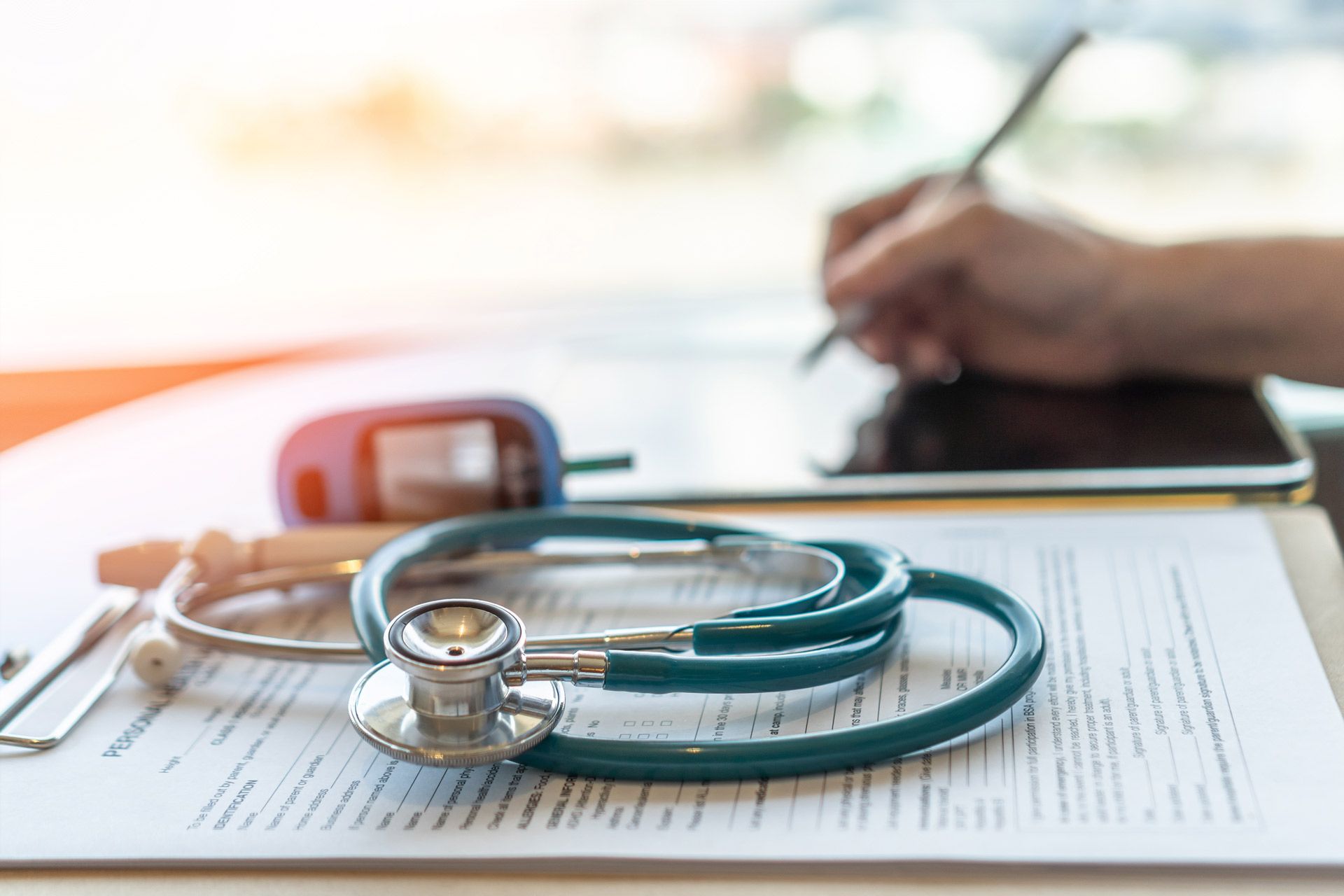 A stethoscope is sitting on top of a clipboard next to a person writing on a tablet