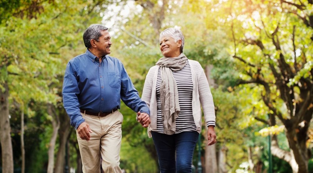 An elderly couple is walking in a park holding hands.