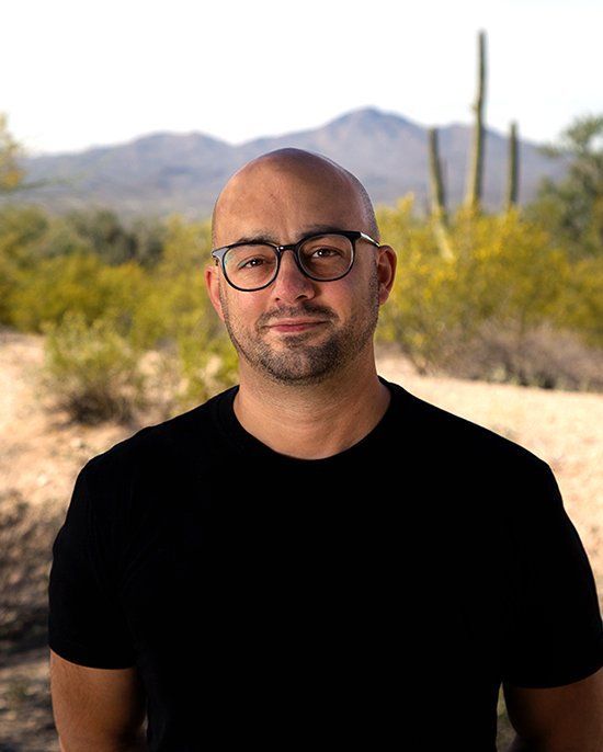 Bald man with glasses smiles, black shirt, in front of desert landscape with mountains.