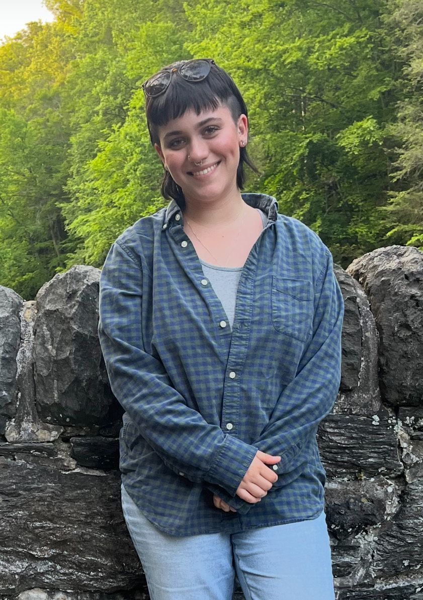 A woman in a plaid shirt is standing in front of a stone wall.