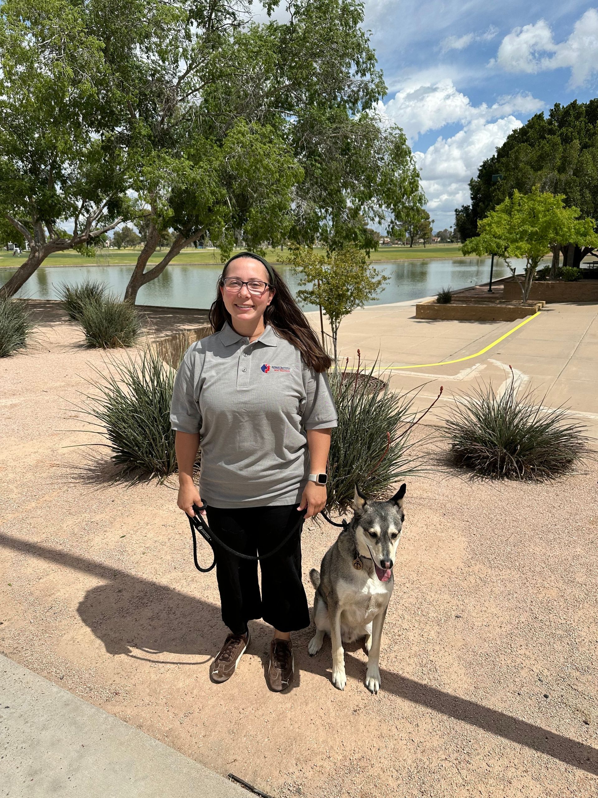 A woman is standing next to a dog on a leash in front of a lake.