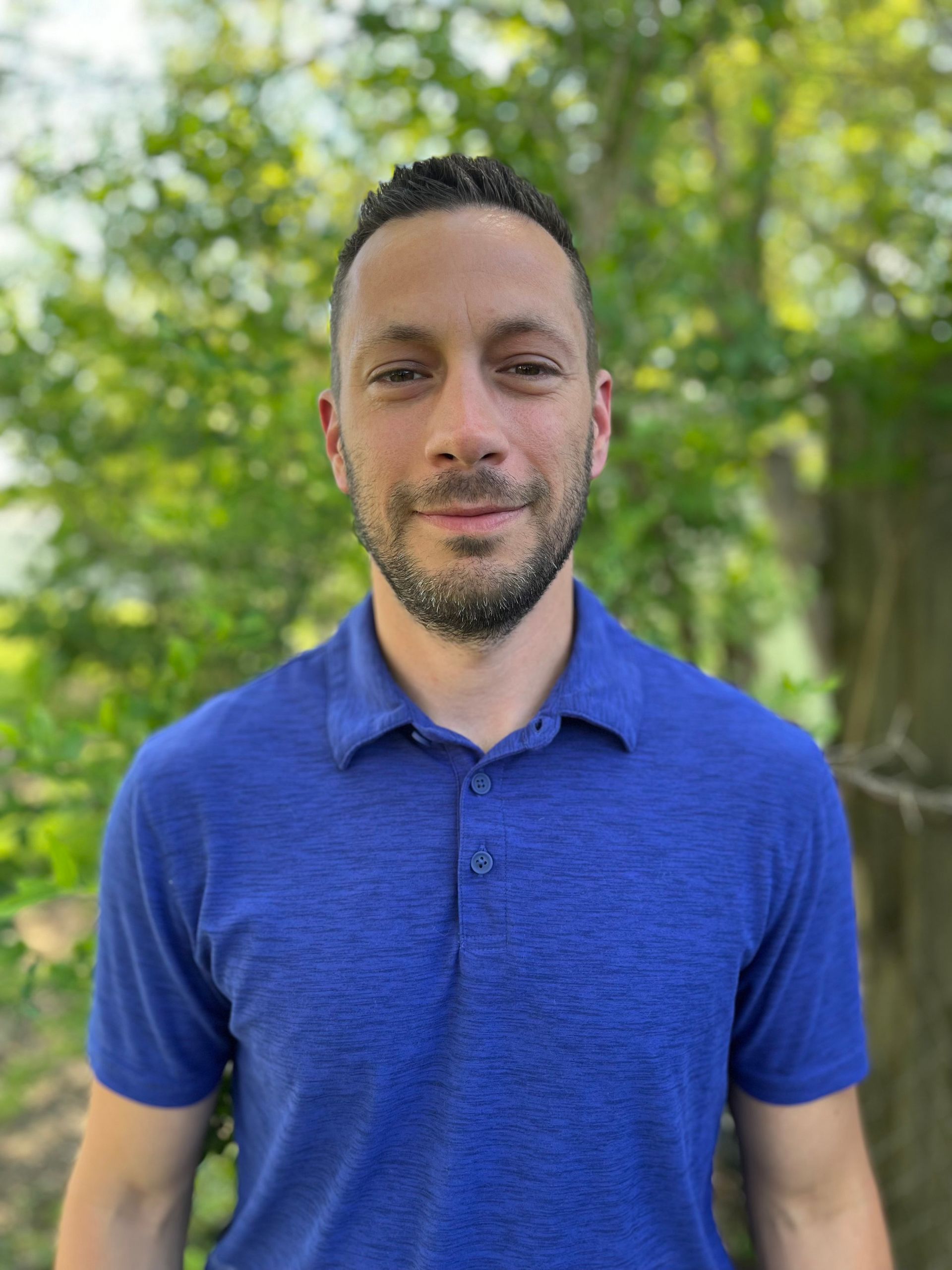 A man with a beard wearing a blue polo shirt is standing in front of trees.