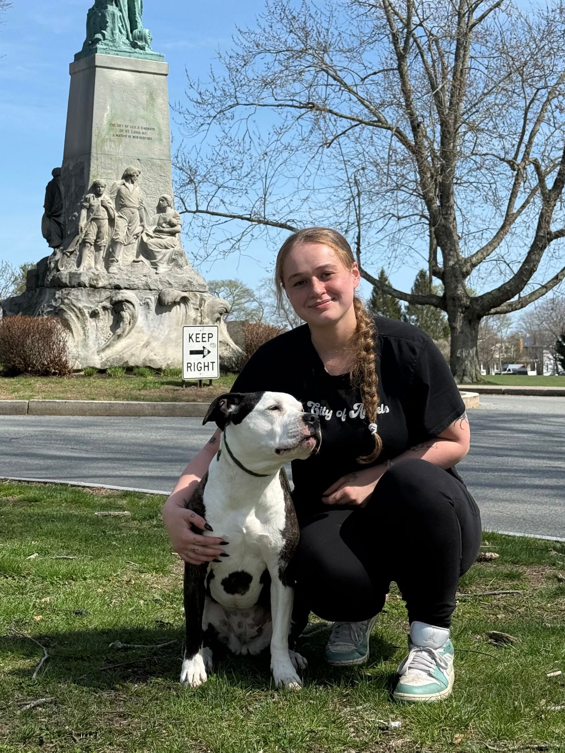 Woman kneels with black-and-white dog in a park, with a monument and bare tree in the background.