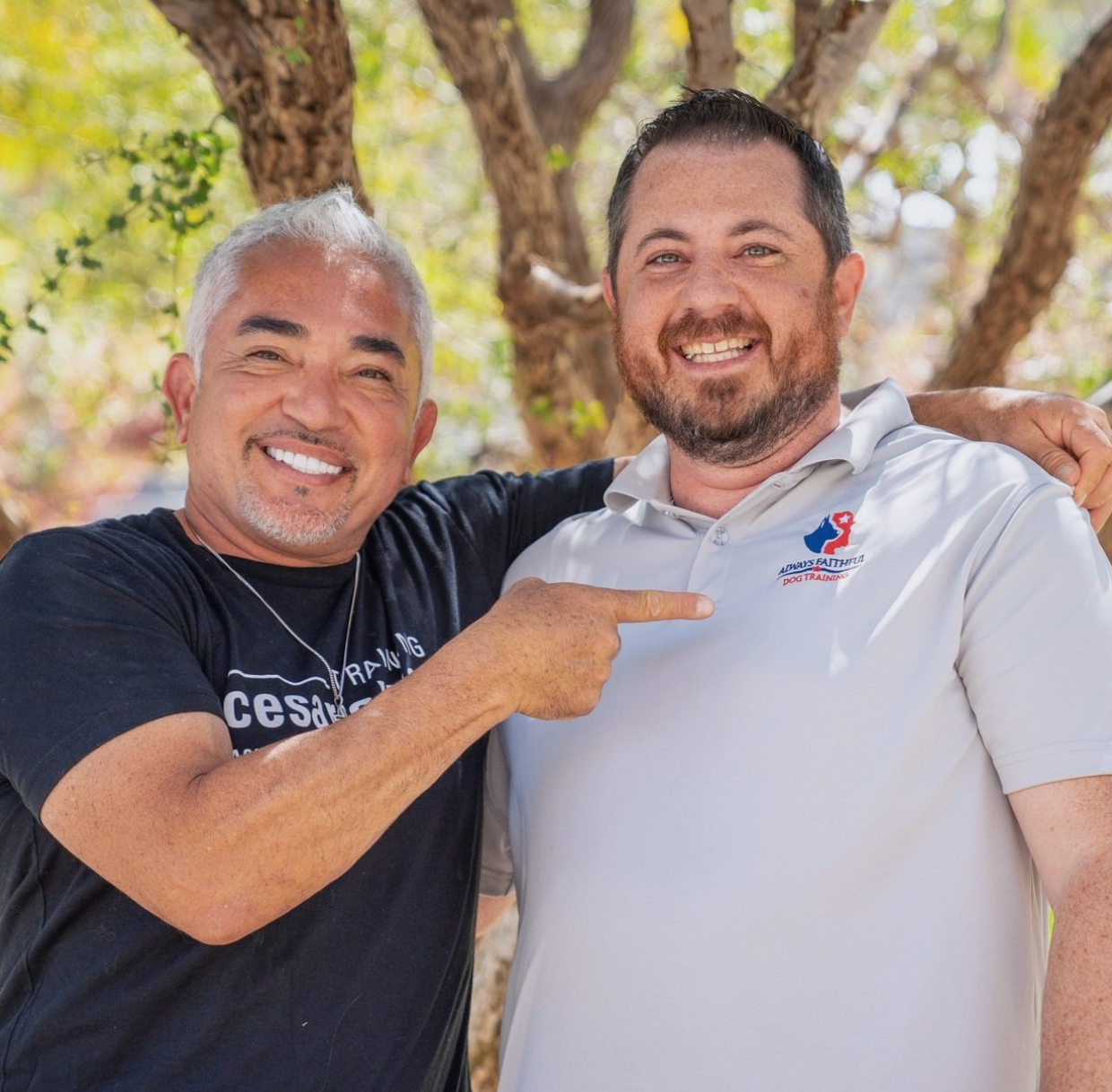 Two men are posing for a picture and one is pointing at his shirt