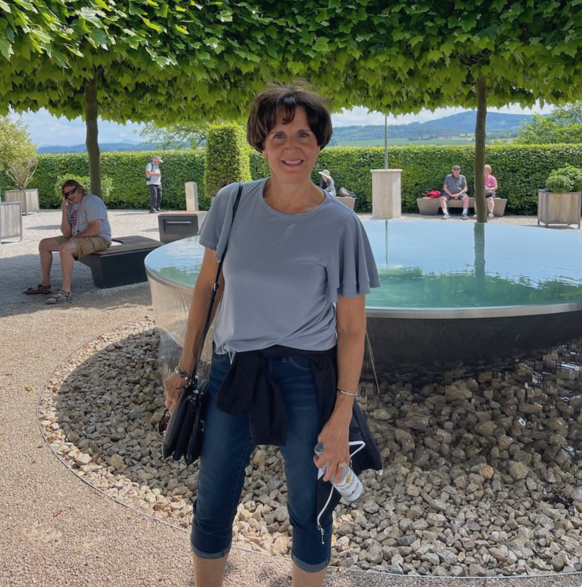 A woman standing in front of a fountain with a man sitting on a bench in the background
