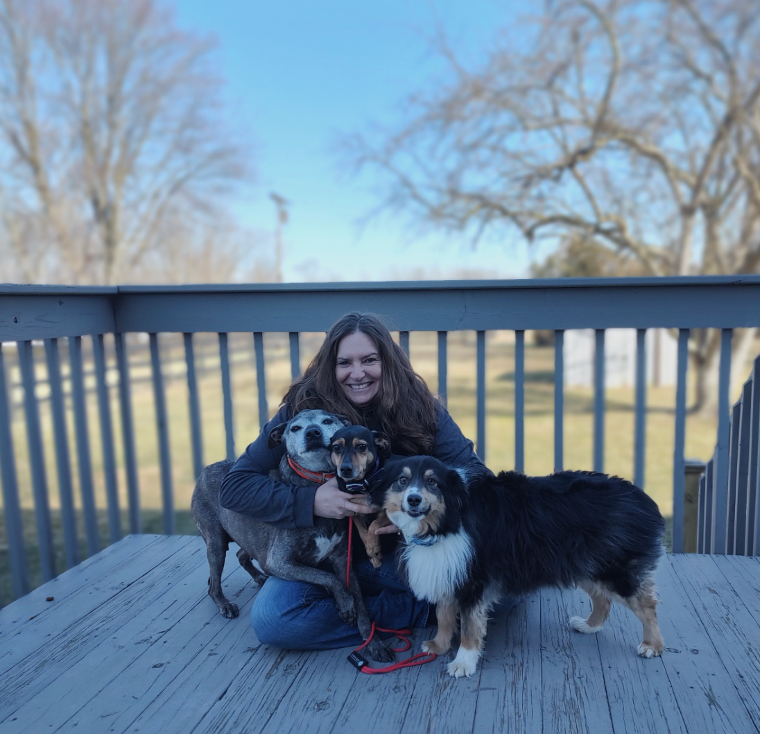 A woman is kneeling down with two dogs on a deck.
