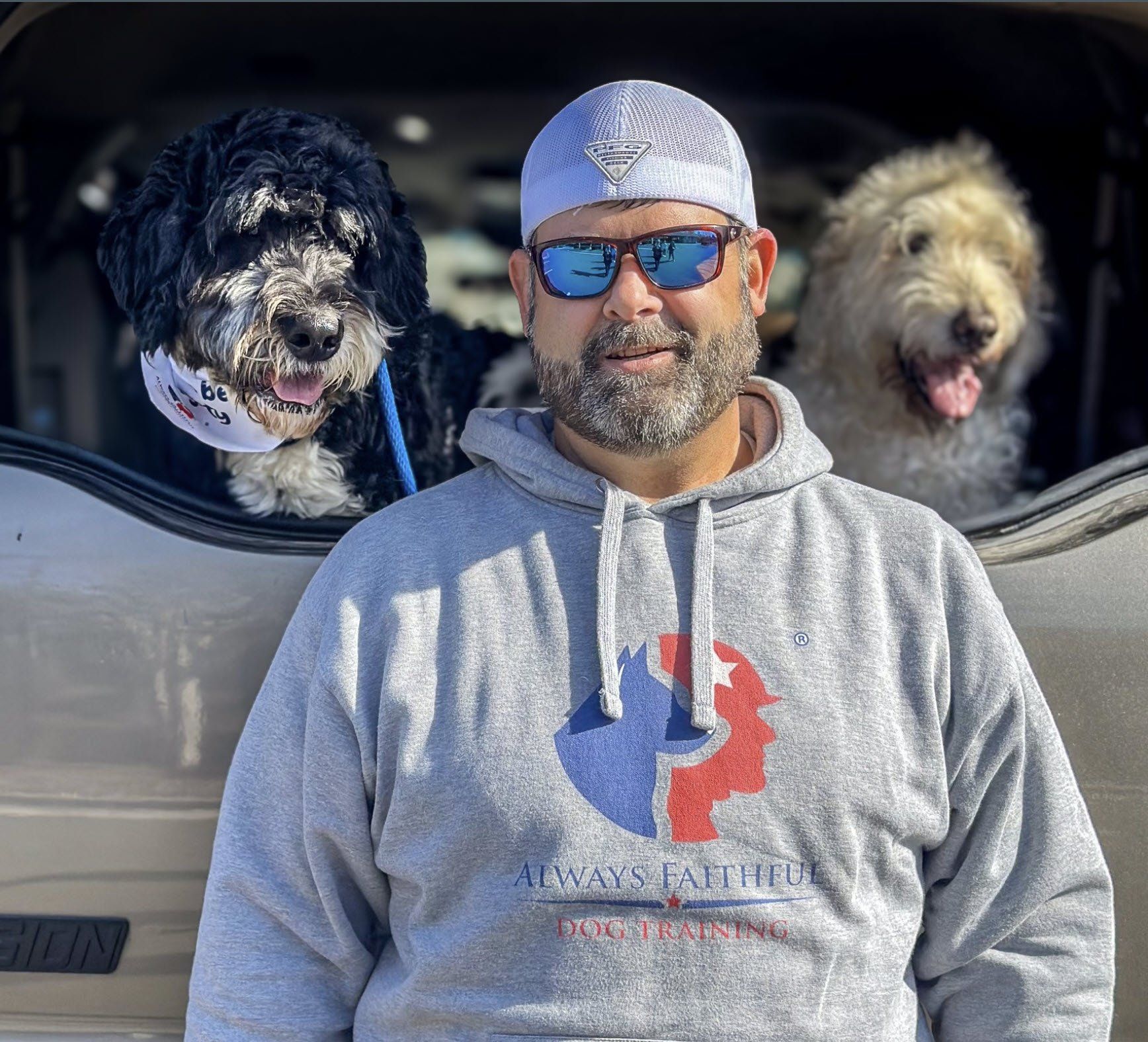 A man wearing sunglasses is standing next to two dogs in the back of a car.