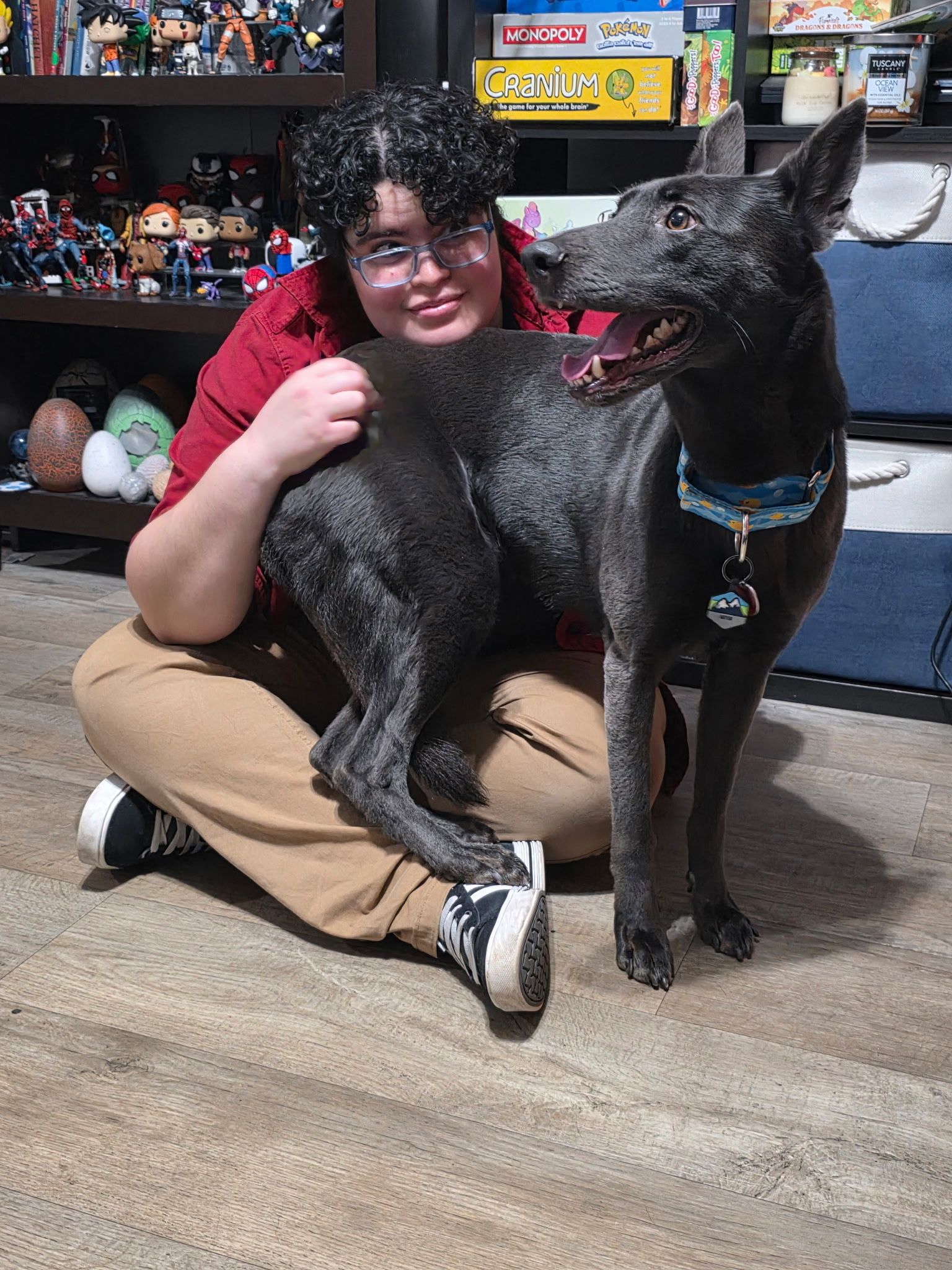 Person with glasses and a black dog seated on the floor, both looking at the camera.