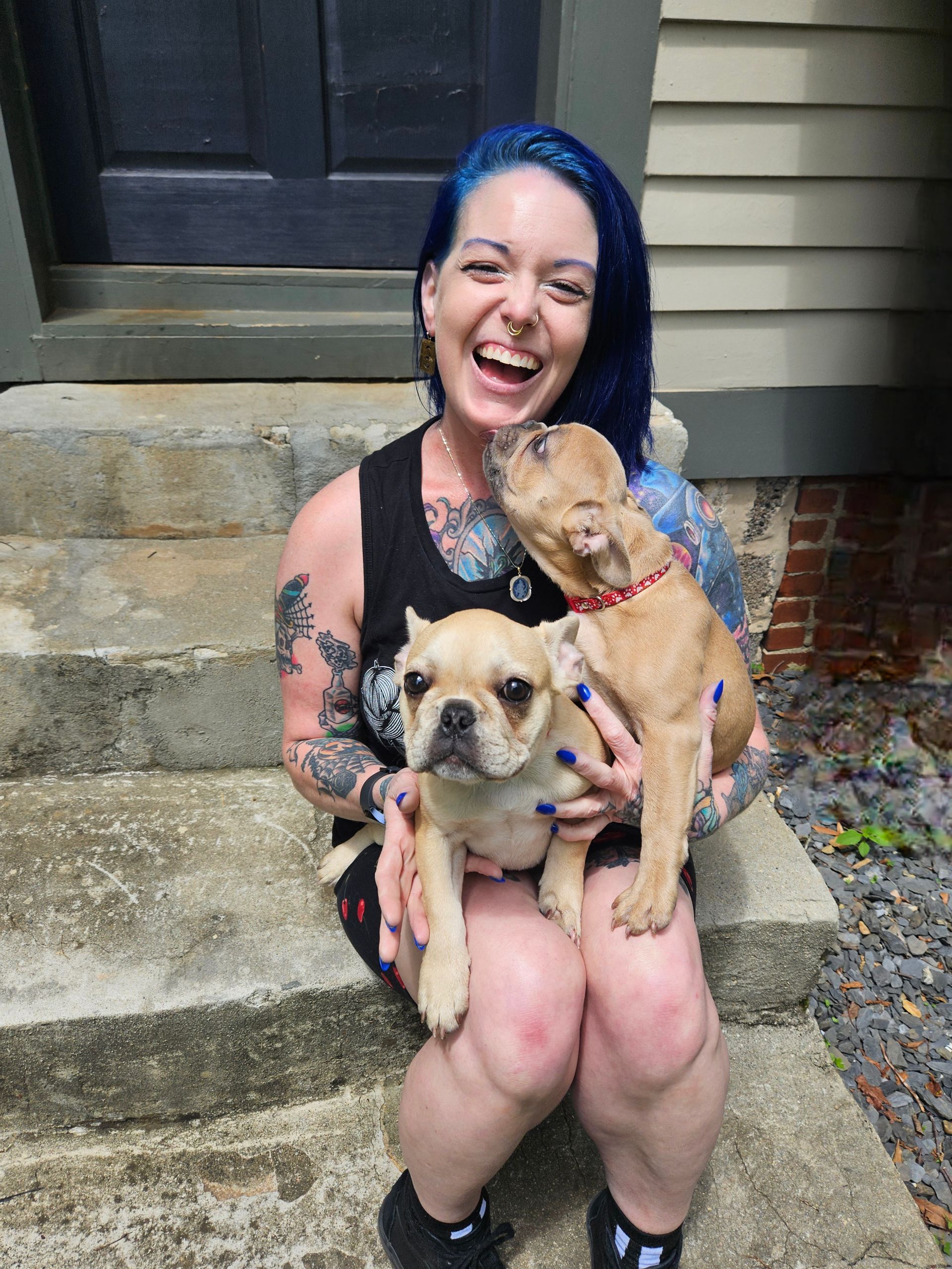 Woman with blue hair and tattoos laughs while holding two French bulldogs on concrete steps.