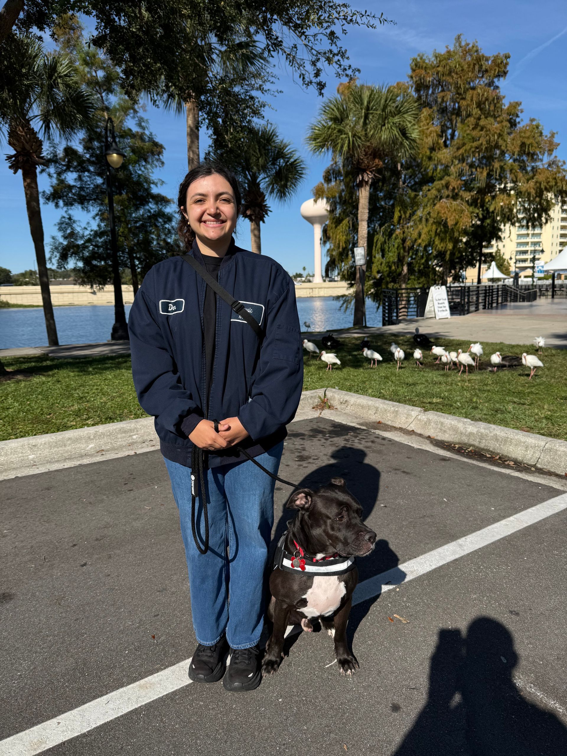 Woman in blue jacket and jeans stands next to a black and white dog on a leash in a parking lot. Water and trees in background.