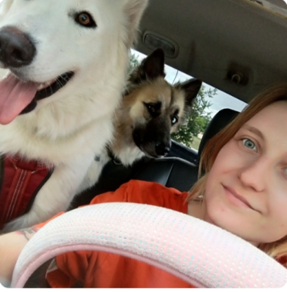 Woman sitting with two dogs in an outdoor setting. The white dog is sitting, the other is lying.