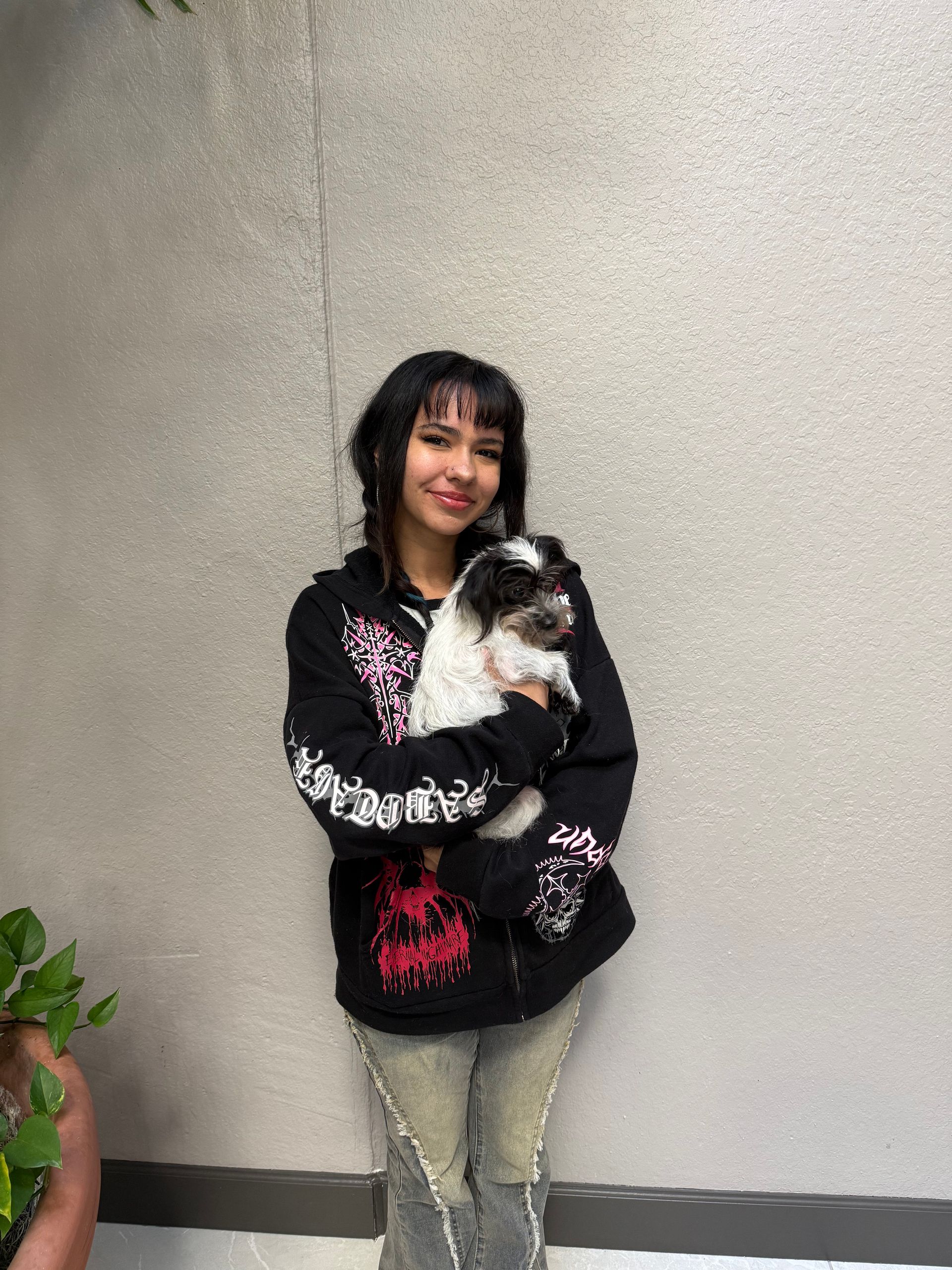 Woman in black hoodie and distressed jeans holding a black and white dog against a gray wall.
