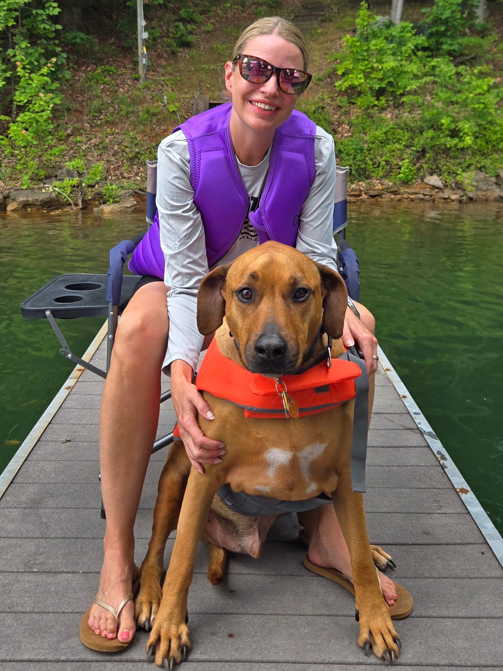A woman is sitting on a dock with a dog wearing a life jacket.