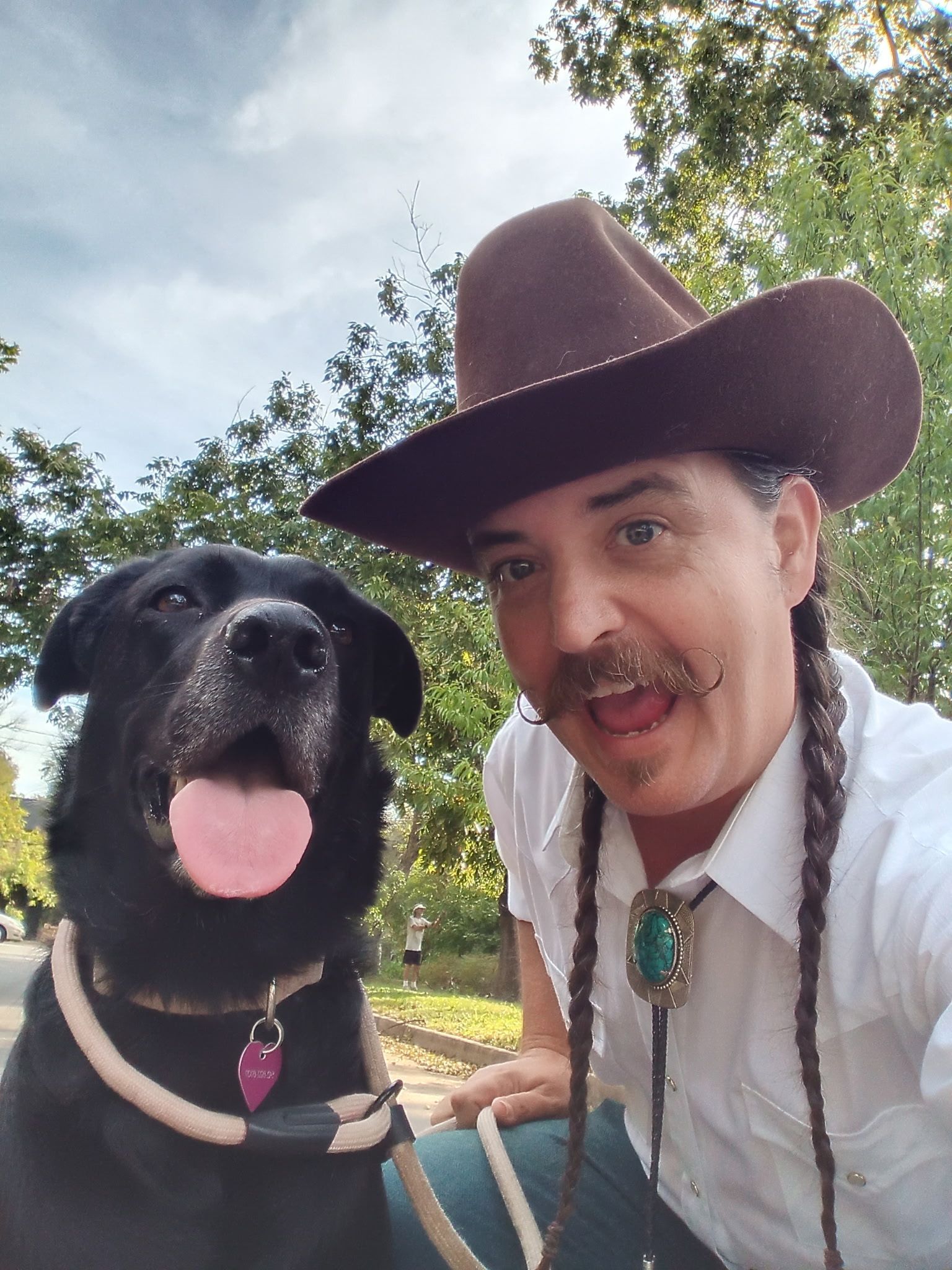 Man in glasses kneels with white dog. Both look towards the camera. Dog’s paw rests on man’s hand. Outdoors, autumn.