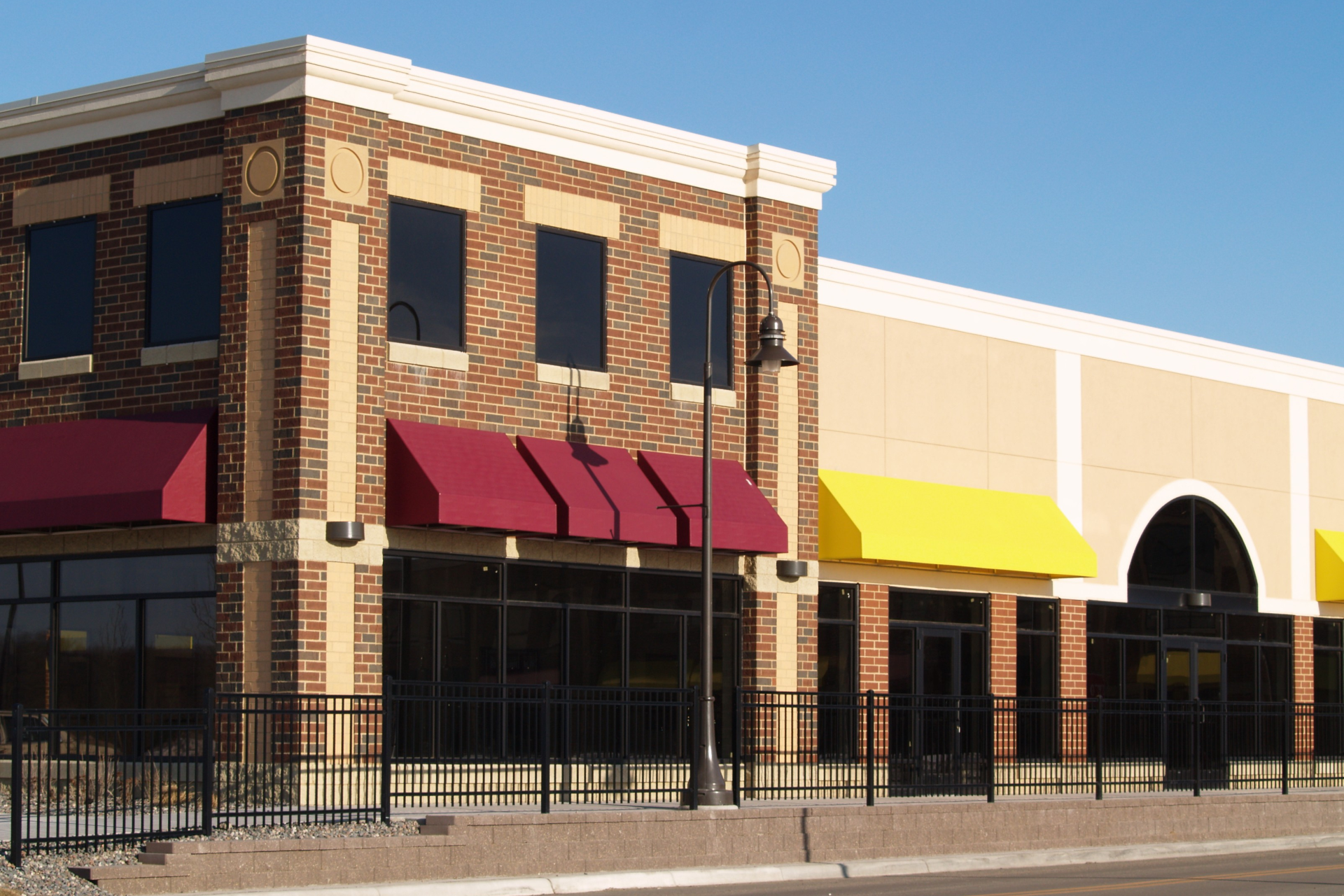 Two-story brick building with red awnings, next to a beige building with a yellow awning, and large windows.