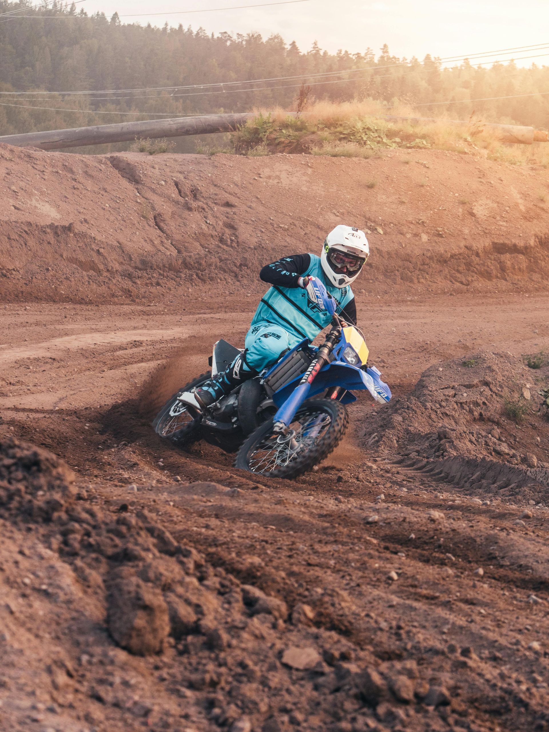 Dirt biker on a blue motorcycle racing around a dirt track, kicking up dust in the sunshine.