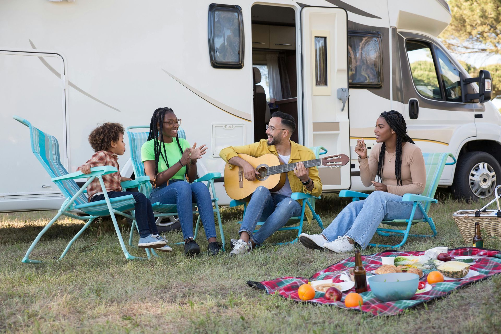 Family sitting outside an RV, one playing guitar, others listening and eating snacks. Picnic blanket.