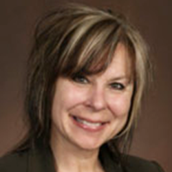 Woman with dark hair and blonde highlights smiles, wearing a dark blazer, against a brown background.