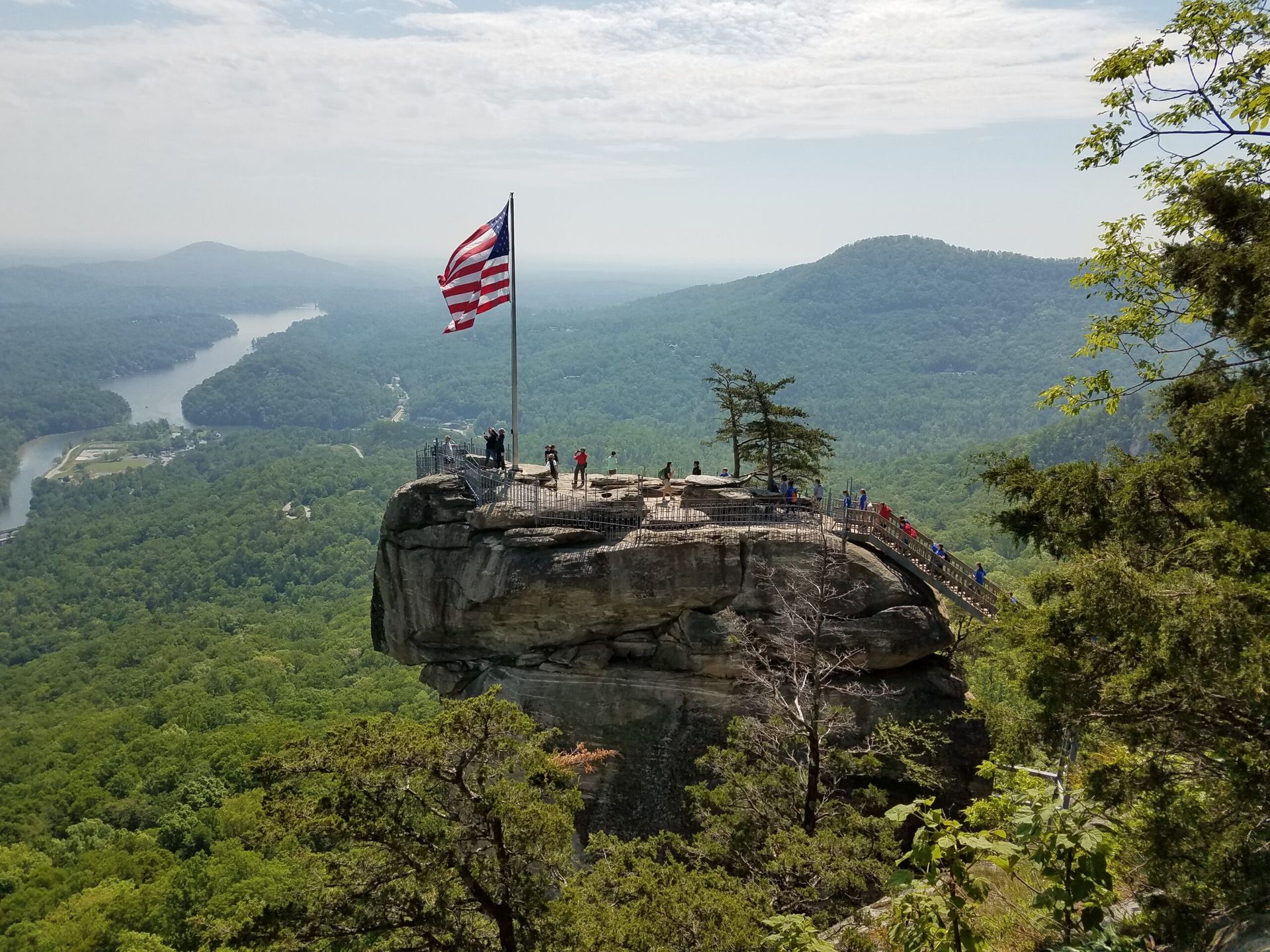 American flag waving on a rocky mountaintop overlook, surrounded by trees and a distant river.