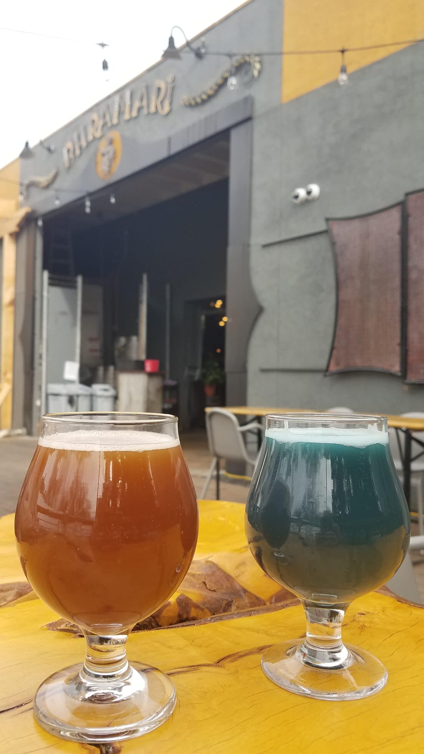Two beers on a yellow table in front of a brewery with open doors. One beer is brown, the other is teal.