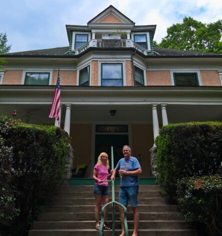 Couple stands on steps of large house with American flag.