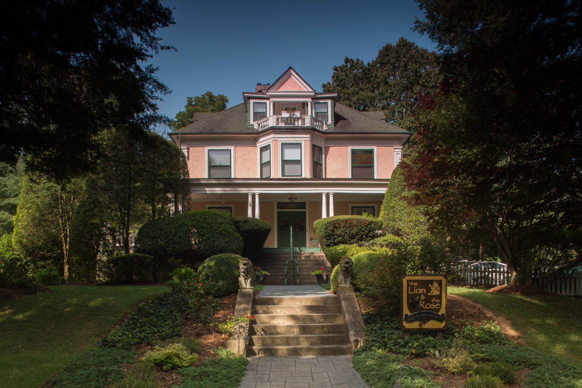 Pink house with white trim, porch, and a staircase leading up to the front door, surrounded by green trees and bushes.