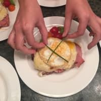 Hands arranging a garnished open-face sandwich with an egg, ham, and tomatoes on a white plate.
