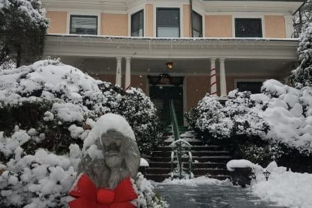 Snow-covered house with porch, shrubs, and steps leading to the door. A red bow decorates a statue in front.