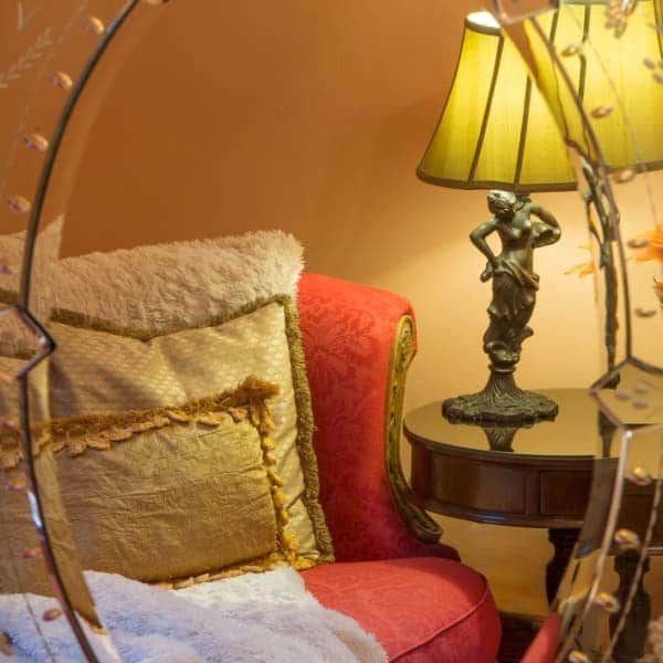 Cozy corner with red chair, pillows, lamp on round table; mirror frames the scene, warm tones.