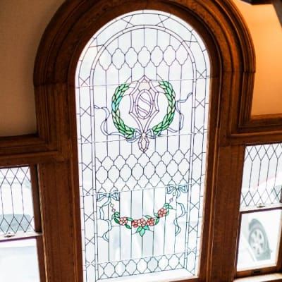 Stained glass window with an arched top, framed in wood. Features a crest, laurel wreath, and floral elements.
