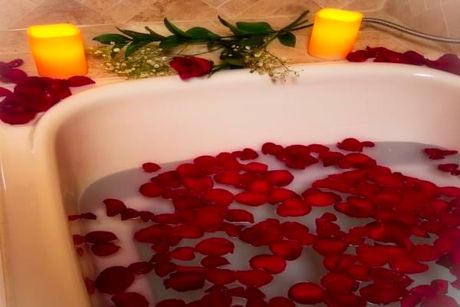 Bathtub filled with milky water and red rose petals; lit candles and greenery on the rim.