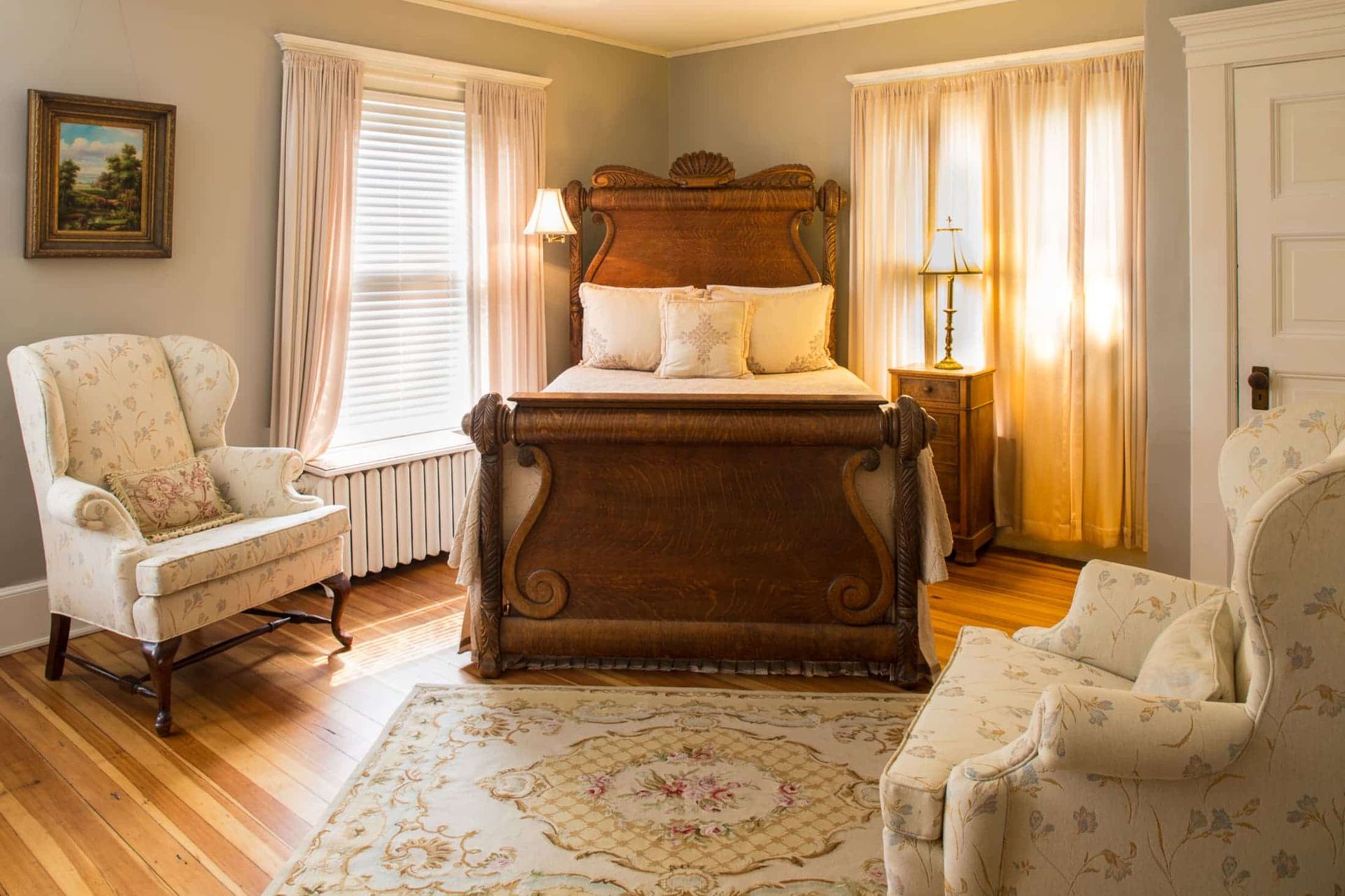 Bedroom with ornate wooden bed, floral patterned armchairs, and soft curtains.