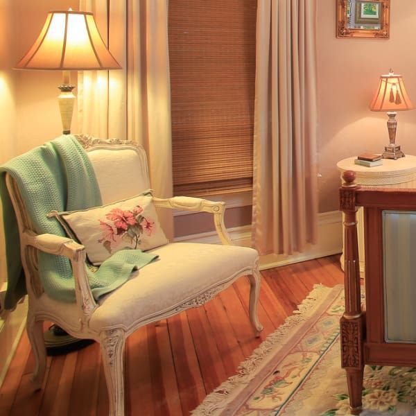 Cozy bedroom with ornate white chair, turquoise throw, floral pillow, and bedside table with lamp. Soft pink walls.