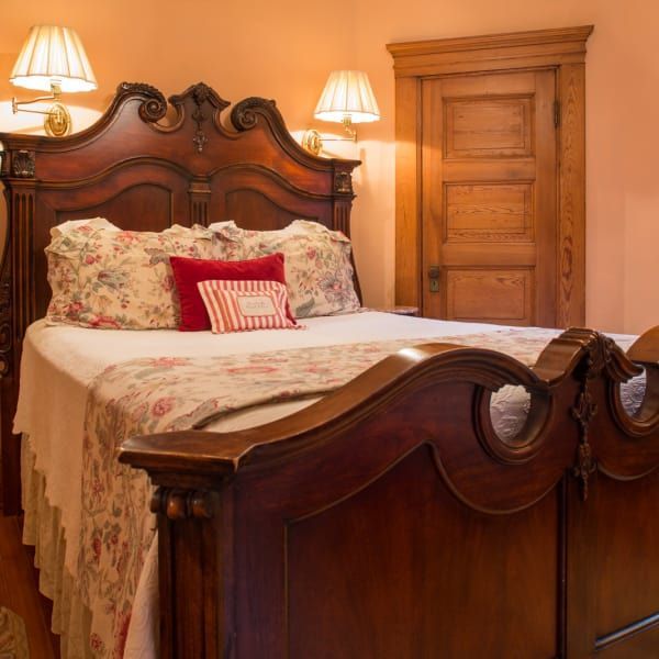 Ornate wooden bed in a bedroom, floral bedding, two wall lamps, closed wooden door, peach walls.
