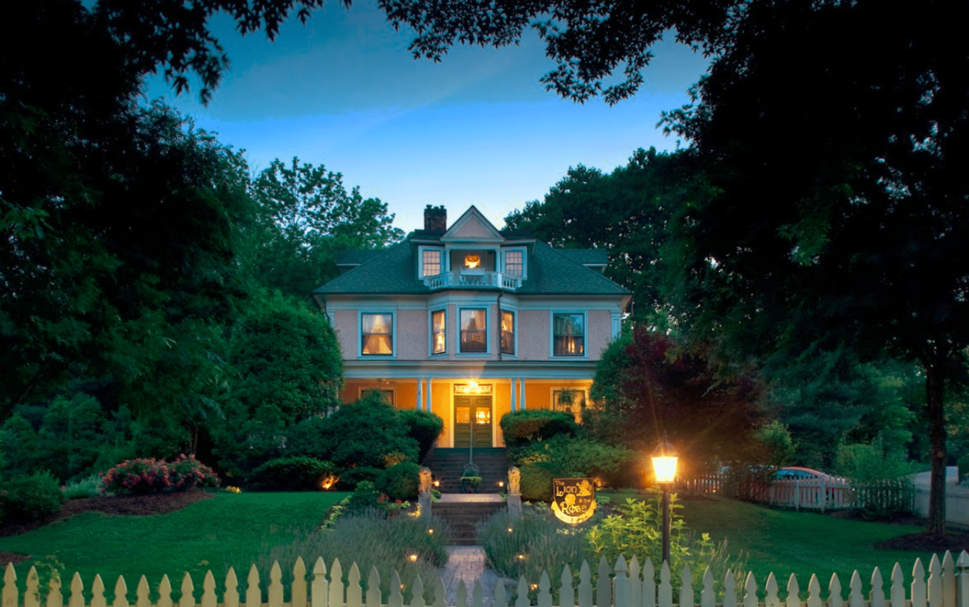Pink house with illuminated windows and porch, surrounded by trees and a white picket fence, at dusk.