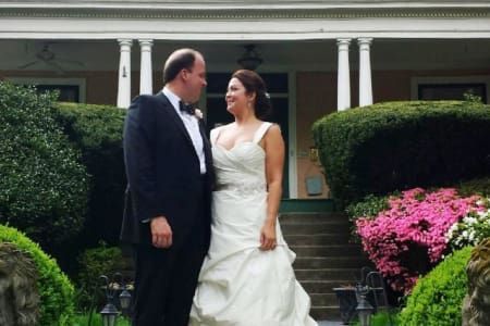 Bride and groom smiling at each other in front of a house. The woman is in a white dress, the man in a black suit.