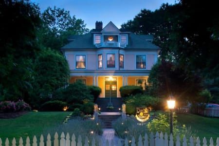 Elegant, multi-story house at dusk, lit from within, with a picket fence and garden.